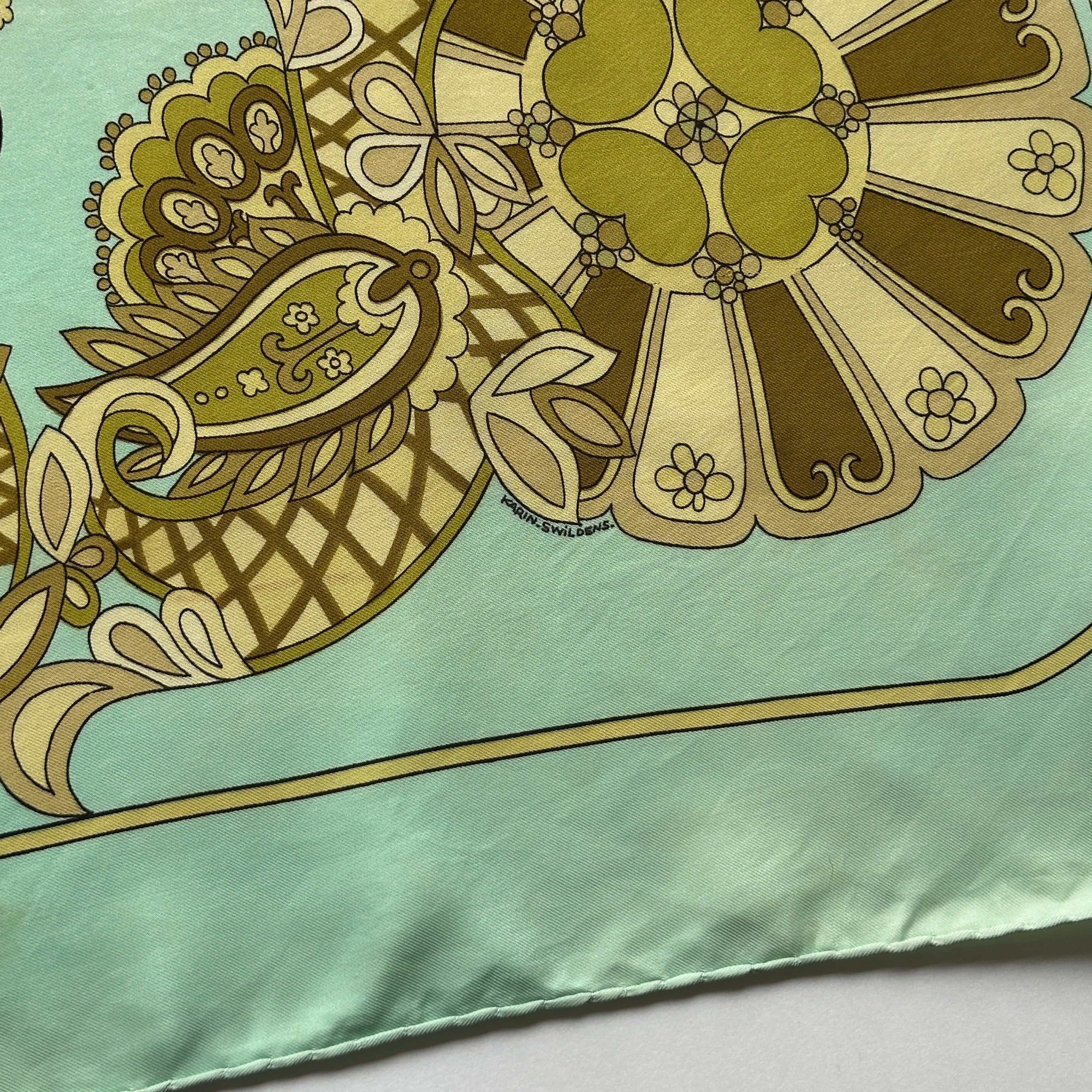 Zoom sur la signature de l'artiste "KARIN SWILDENS" intégrée dans un motif floral géométrique vert et brun.