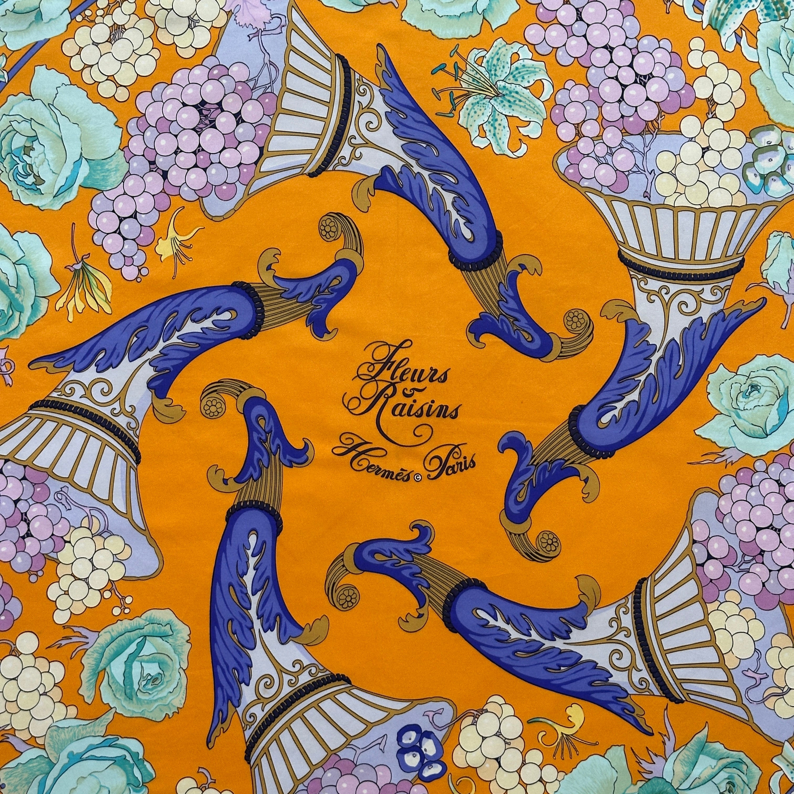 Zoom sur le centre du foulard jaune safran montrant la calligraphie "Fleurs & Raisins - Hermès Paris" entourée de motifs baroques bleus.