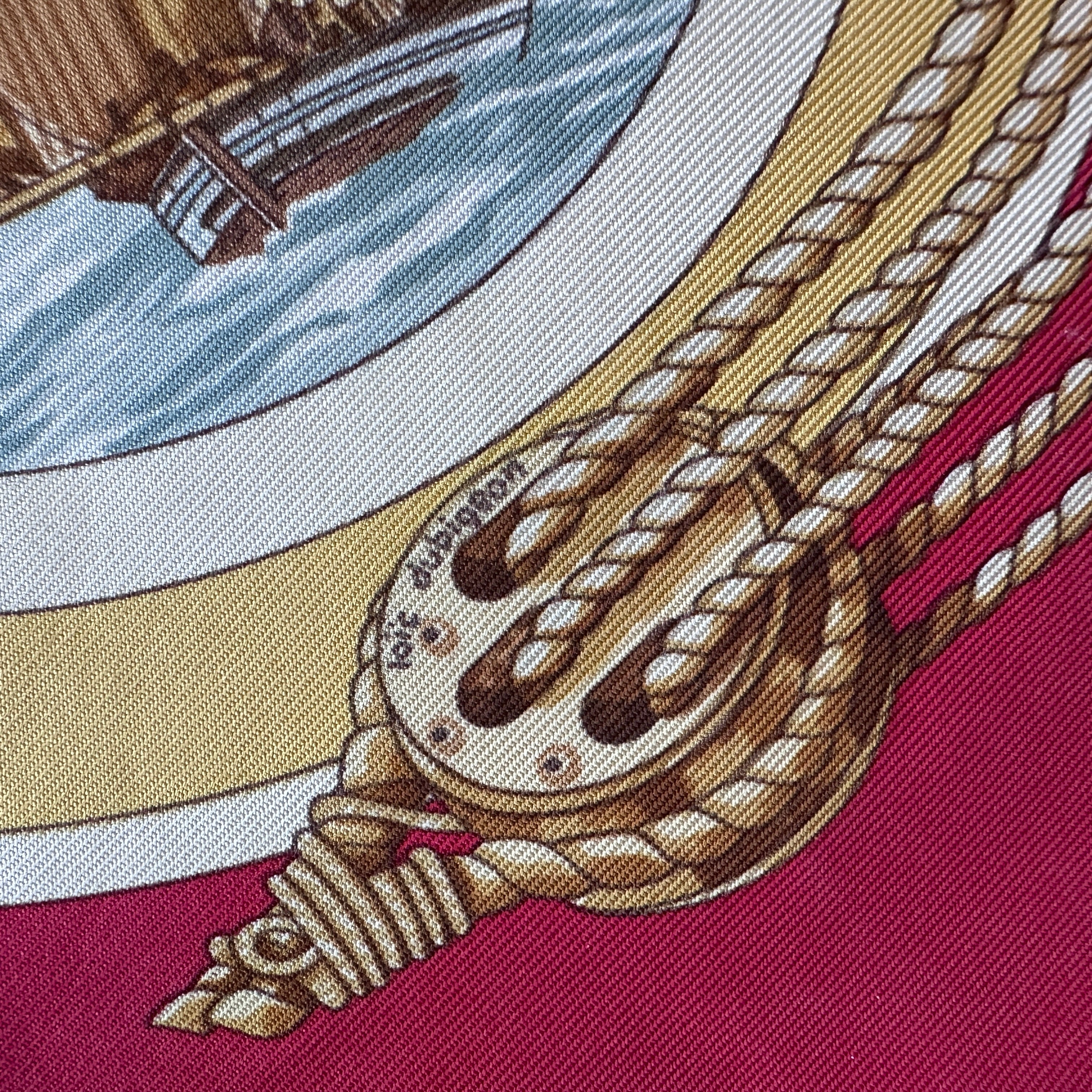 Gros plan sur une poulie et cordages dorés sur fond rouge Hermès avec signature loic dubigeon dans le dessin